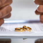 two hands from both sides of a table on top of divorce paperwork and two wedding rings