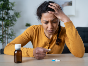 A distraught woman sits in front of a pile of pills with her head in her hand.
